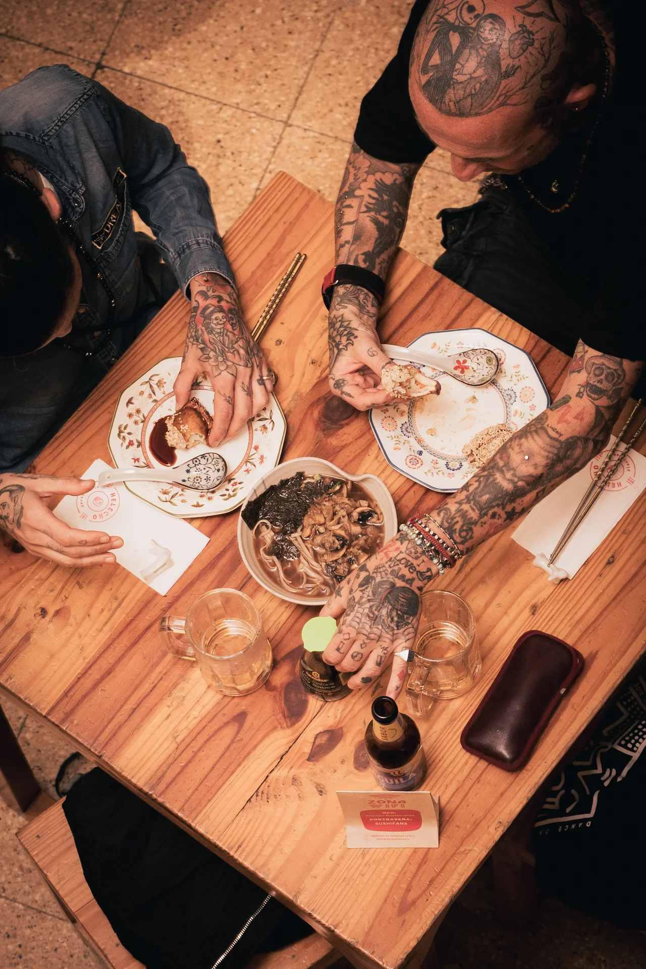 Fotografía gastronómica cenital de comensales con tatuajes comiendo ramen y sushi en mesa de madera, restaurante plant based — Jose Fonseca Medellín