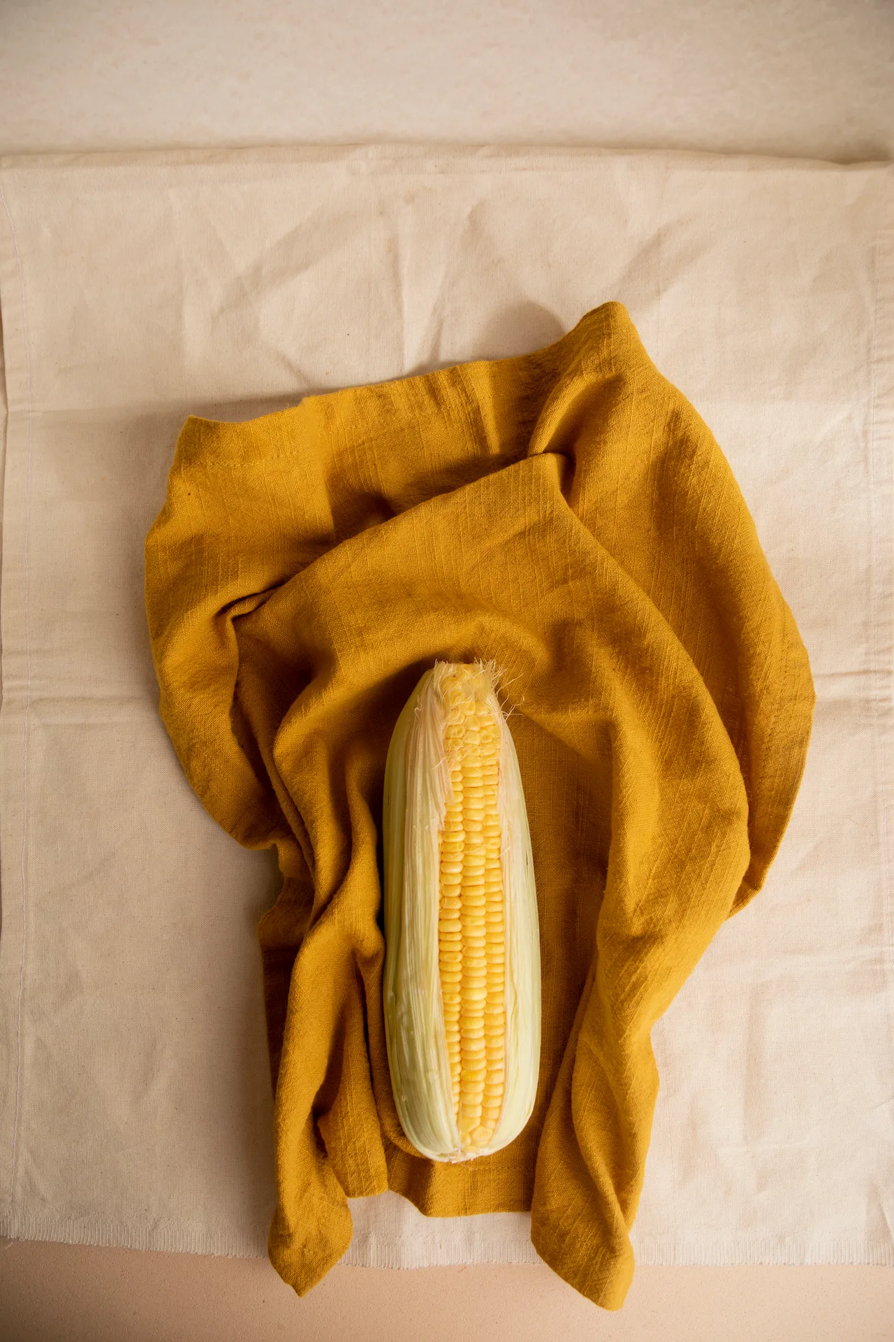 Fotografía gastronómica de mazorca fresca parcialmente pelada sobre tela de lino mostaza, vista cenital — Jose Fonseca Medellín